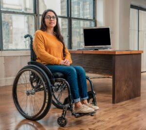 žena na vozíku sedí v kanceláři u pracovního stolu, na kterém je počítač